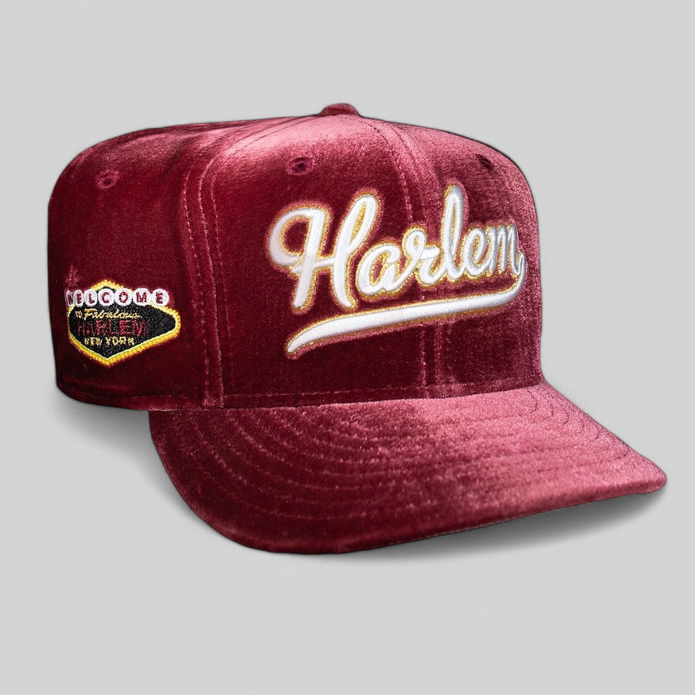 A red fitted baseball cap with 'Harlem' written on it in white script, featuring a grey bottom and MLB branding on the side.